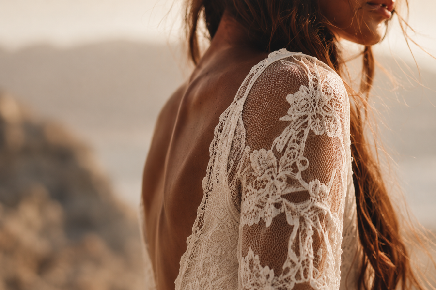 A close-up lifestyle shot of a woman wearing a white lace bridal top or dress, showing the intricate floral lace pattern on her shoulder and back, set against a warm, blurred outdoor background.